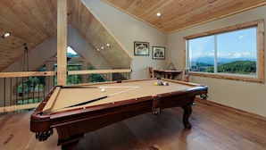 Una mesa de billar en una sala con pisos de madera y un techo alto, una ventana con vista a la montaña, y una escalera que conduce a un nivel superior.