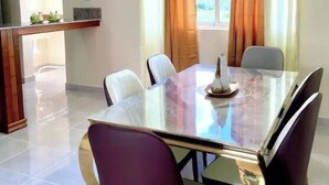 Un coin repas avec une table et des chaises en verre, une cuisine à l’arrière-plan et des rideaux aux fenêtres.