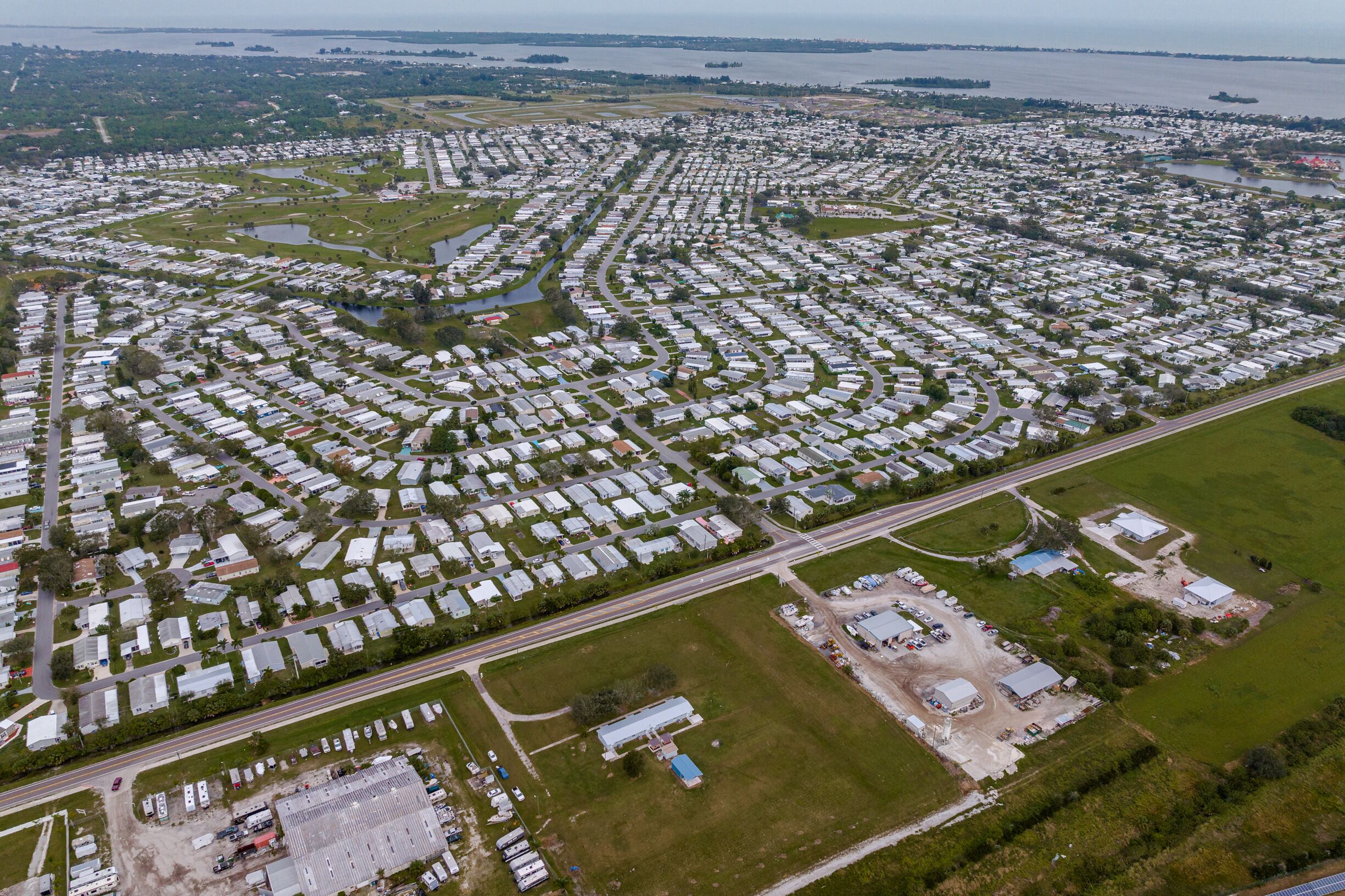 An aerial view of a large campground with numerous RVs and trailers arranged in a grid pattern, surrounded by green spaces and a body of water in the background.