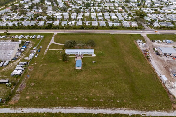 An aerial view of a residential area with a large open grassy field, a small building with a blue roof, and a row of mobile homes in the background.