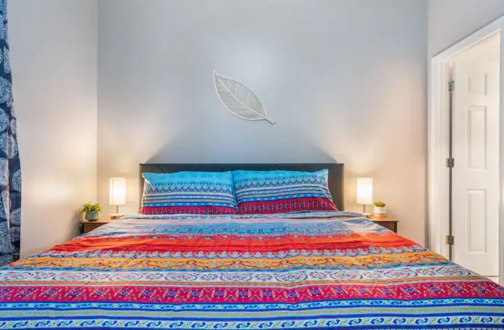 Un lit avec une couvre-lit et des coussins aux motifs colorés, un mur blanc orné d’un motif de feuilles et deux lampes de chevet.