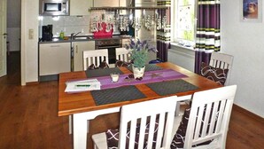 Un coin repas avec une table en bois, des chaises blanches, un vase avec des fleurs et, au fond, une cuisine.