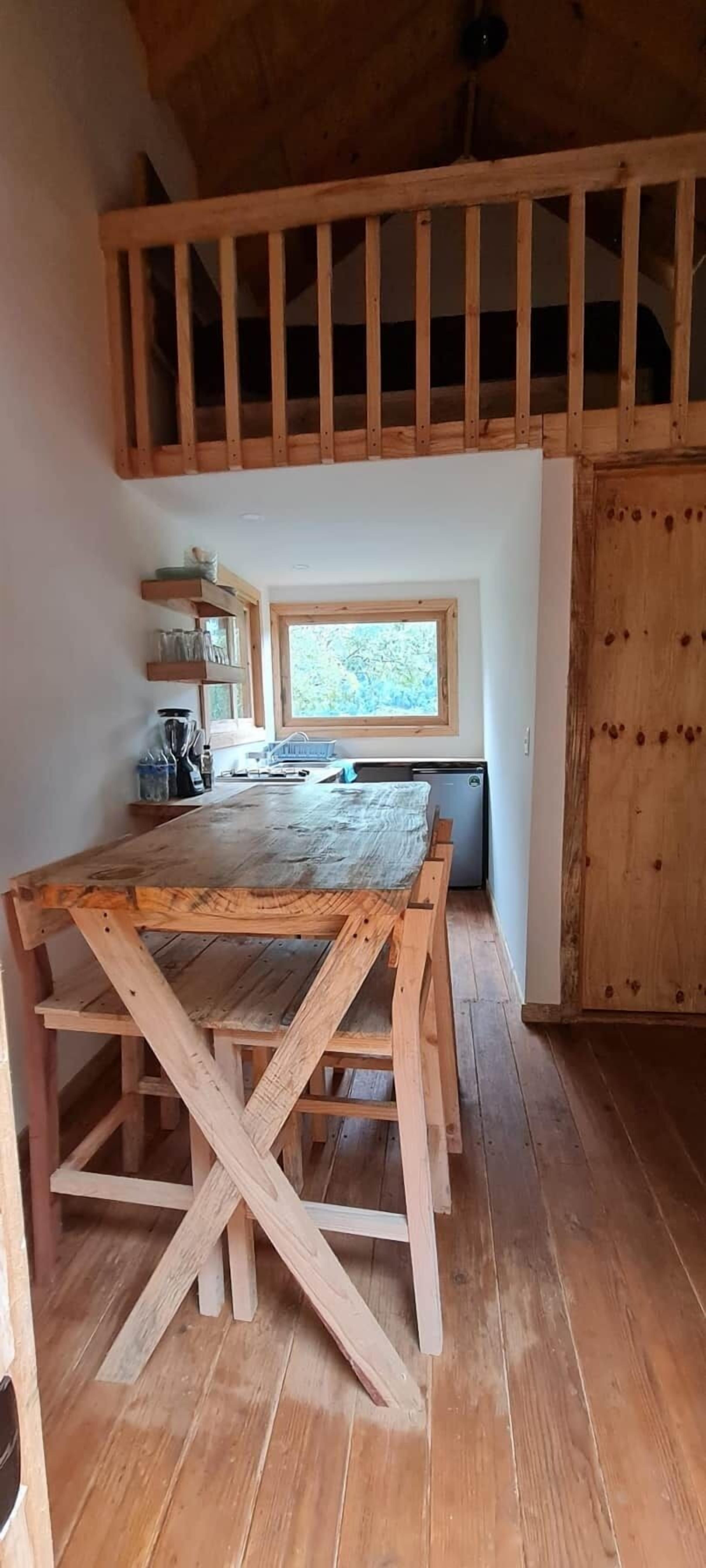 Et rustikt træspisebord med bænke, et vindue med udsigt til grønne omgivelser, og et træloft.