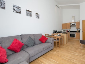 Uma sala de estar moderna com um sofá cinzento, almofadas vermelhas e cinzentas, e uma mesa de jantar de madeira. Cozinha visível ao fundo, com armários de madeira e um fogão.