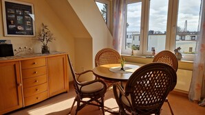 A dining area with a round table and wicker chairs, a wooden cabinet with drawers, and a window with a view of buildings and a tower. - Ferienwohnung, Grömitz (Grömitz)