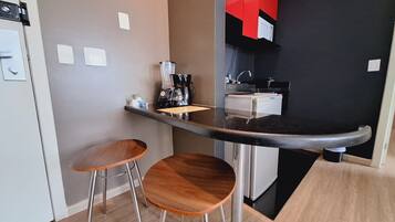 Uma cozinha compacta equipada com cafeteira, mini-geladeira e dois banquinhos de madeira.