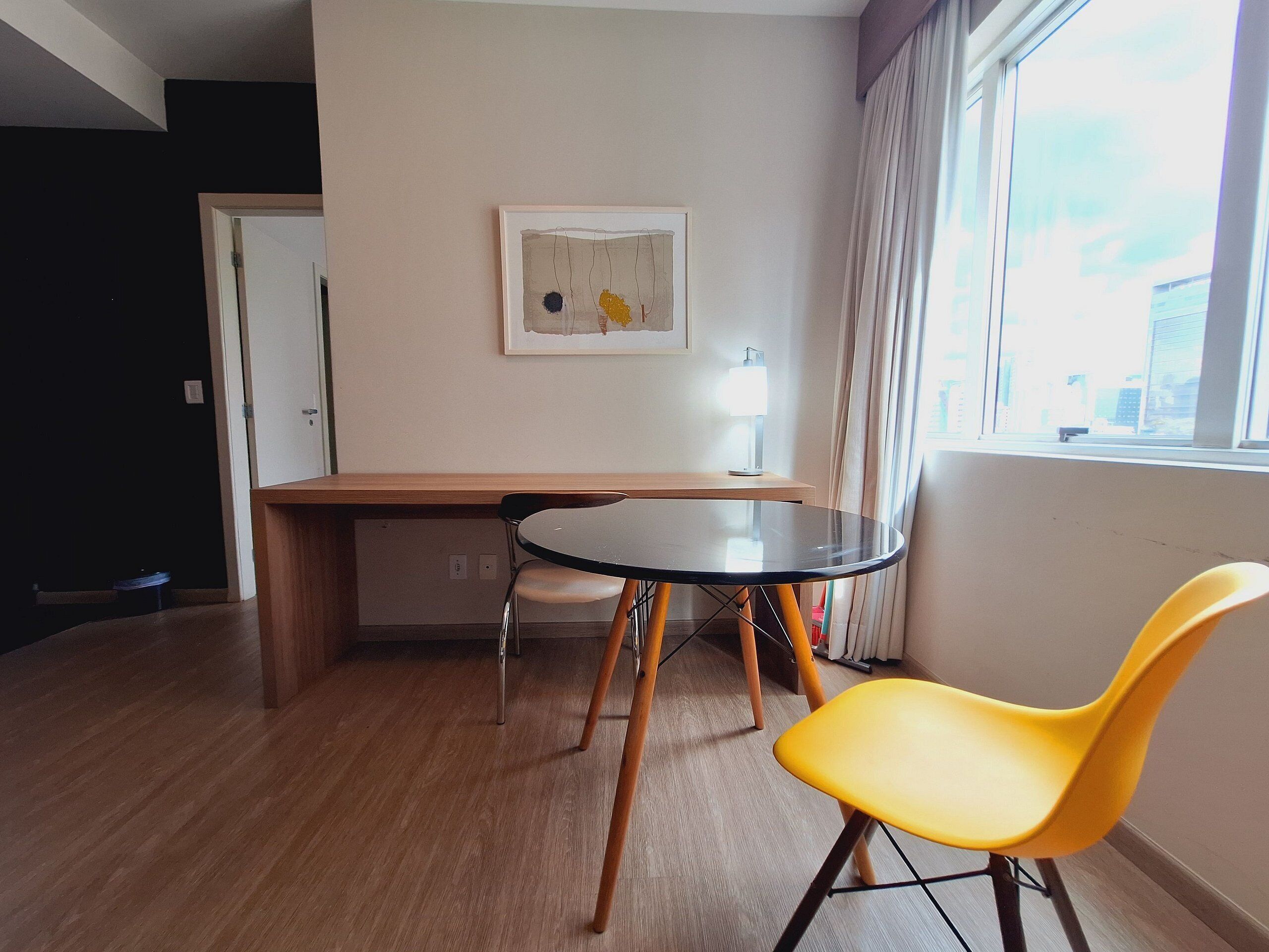 Una habitación con piso de madera, una mesa redonda con sillas, un cuadro enmarcado en la pared y una ventana con cortinas.