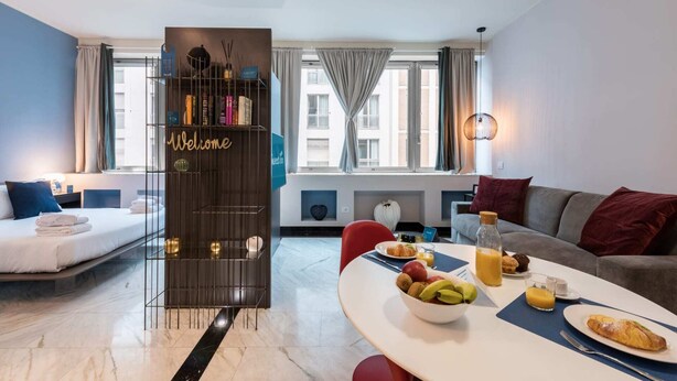 Une chambre d’hôtel moderne équipée d’un lit, d’un canapé, d’une table à manger avec un petit-déjeuner, d’une bibliothèque et offrant une vue sur la ville grâce à de grandes fenêtres.
