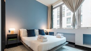 Une chambre d’hôtel moderne, équipée d’un lit, de tables de chevet, d’une fenêtre avec des rideaux et offrant une vue sur les bâtiments environnants.