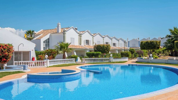 A swimming pool with a tiled edge, surrounded by white buildings with red-tiled roofs, and a clear blue sky.