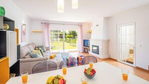 A bright living room with a sofa, coffee table, and a dining table with fruit and drinks. There is a TV, a fireplace, and a sliding glass door to a garden.