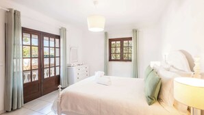 Een slaapkamer met een bed, nachtkastje, lamp, dressoir en een raam met gordijnen.