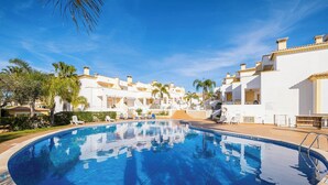 A swimming pool surrounded by white buildings with balconies, palm trees, and a clear blue sky.
