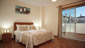 A bedroom with a large bed, a nightstand, a painting on the wall, and a sliding glass door to the outside.