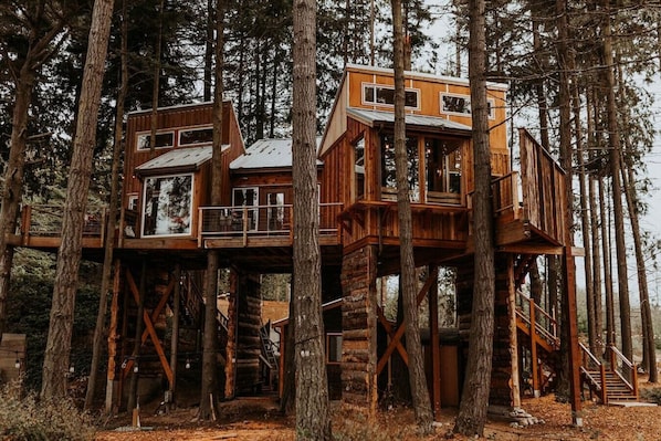 Ein hölzernes Baumhaus mit mehreren Ebenen, das auf Stelzen steht und von hohen Bäumen umgeben ist.