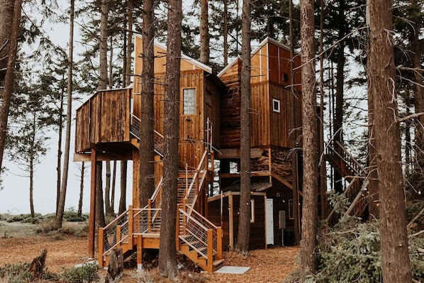 A wooden treehouse with multiple levels, built around a central tree, featuring a staircase and a small door.