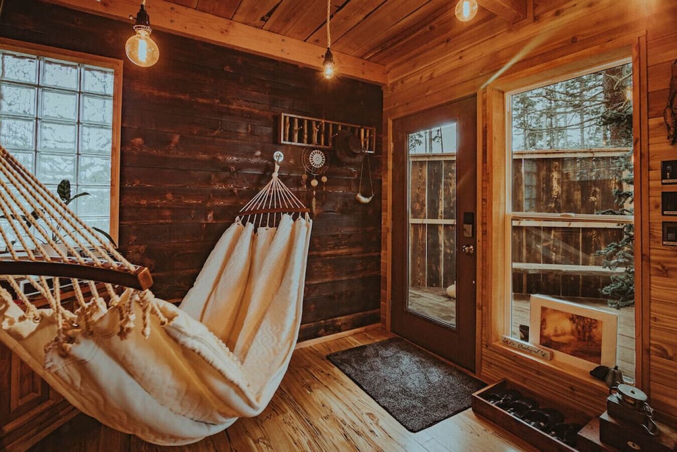 A wooden cabin interior with a hammock, a wooden door, and a view of trees outside.