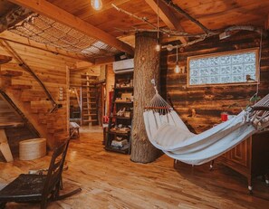 A rustic cabin interior with a hammock, wooden floors, a staircase, and a bookshelf.