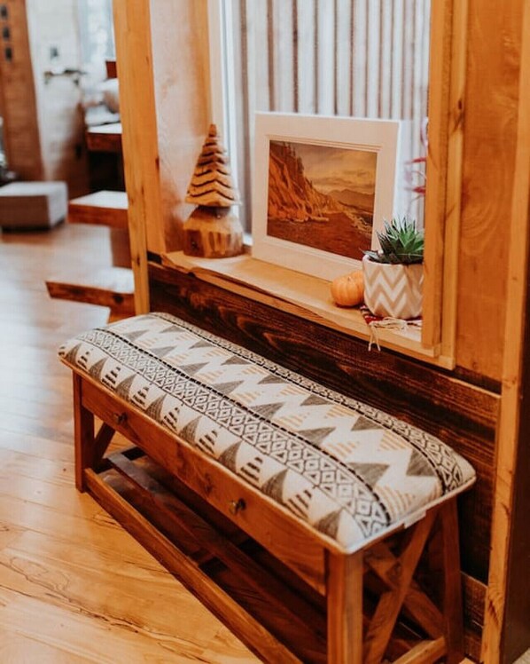 A wooden bench with a patterned cushion, a small pine tree decoration, and a framed picture on a wooden shelf.