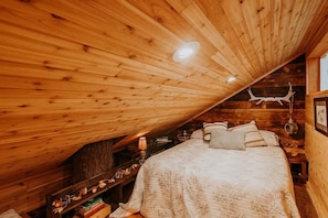 Ein gemütliches Schlafzimmer im Dachgeschoss, mit schrägen Wänden, einem Bett mit vielen Kissen, einem kleinen Tisch mit Lampe sowie einem Regal mit Büchern und Dekorationen.