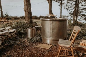 A rustic outdoor setting with a large metal barrel, a smaller metal container, and a wooden chair.