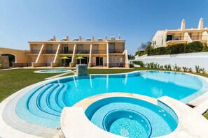 A swimming pool with a hot tub, surrounded by a well-maintained lawn and a building with multiple balconies.