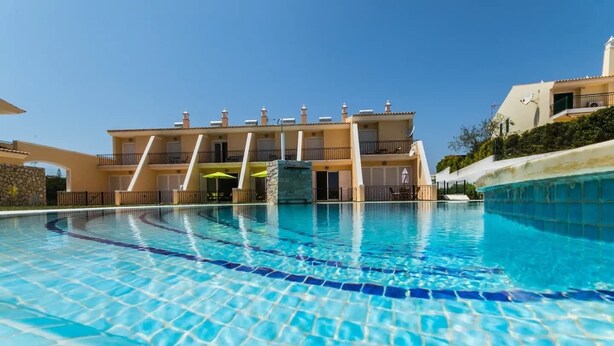 A swimming pool with a tiled floor, surrounded by a building with balconies and a stone wall. - Villa Filipa III - Beachfront with Jacuzzi (Albufeira)