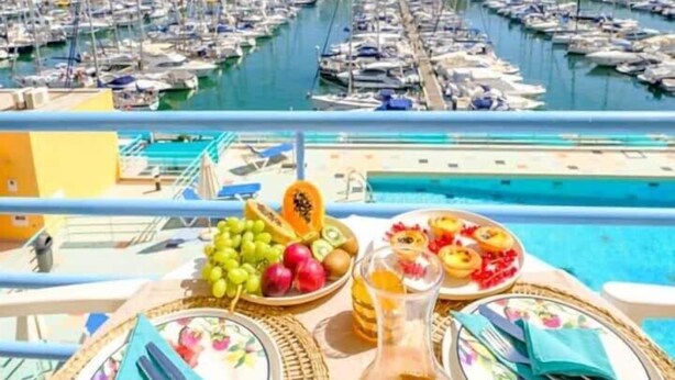 Mesa de comedor con platos, cubiertos y una botella de jugo de naranja, con vista a un puerto deportivo y barcos.