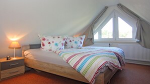 Ett sovrum med en säng i trä, blommig dekorerade kuddar, en randig filt, ett nattduksbord med en lampa och ett fönster med gardiner.