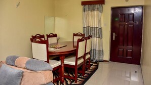 A dining area with a wooden table and chairs, a sofa, a patterned rug, a door, and a ceiling light.