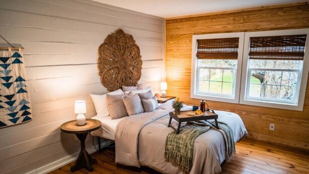A bedroom with a wooden floor, a bed with a decorative headboard, a bedside table with a lamp, and a window with blinds.