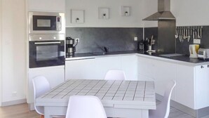 Cozinha moderna com armários brancos, bancada preta, coifa em aço inoxidável e área de jantar com mesa e cadeiras brancas.