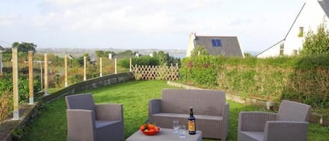 A patio with wicker furniture, a table with a bottle and glasses, and a view of a white building with a sloped roof.