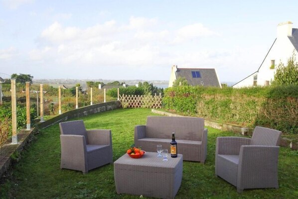 A patio with wicker furniture, a table with a bottle and glasses, and a view of a white building with a sloped roof.