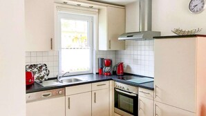 Eine kleine Küche mit weißen Schränken, einer schwarzen Arbeitsplatte, einer Edelstahl-Dunstabzugshaube, einer roten Kaffeemaschine und einem Fenster mit Jalousien.