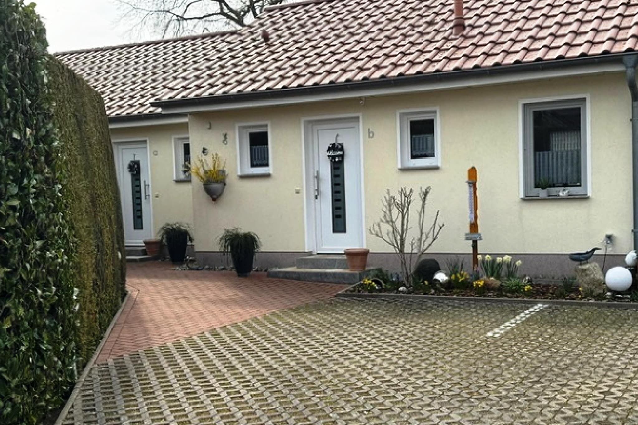 Un bâtiment avec un toit recouvert de tuiles, une porte blanche et un petit jardin agrémenté de plantes en pot et d’un abreuvoir pour oiseaux.