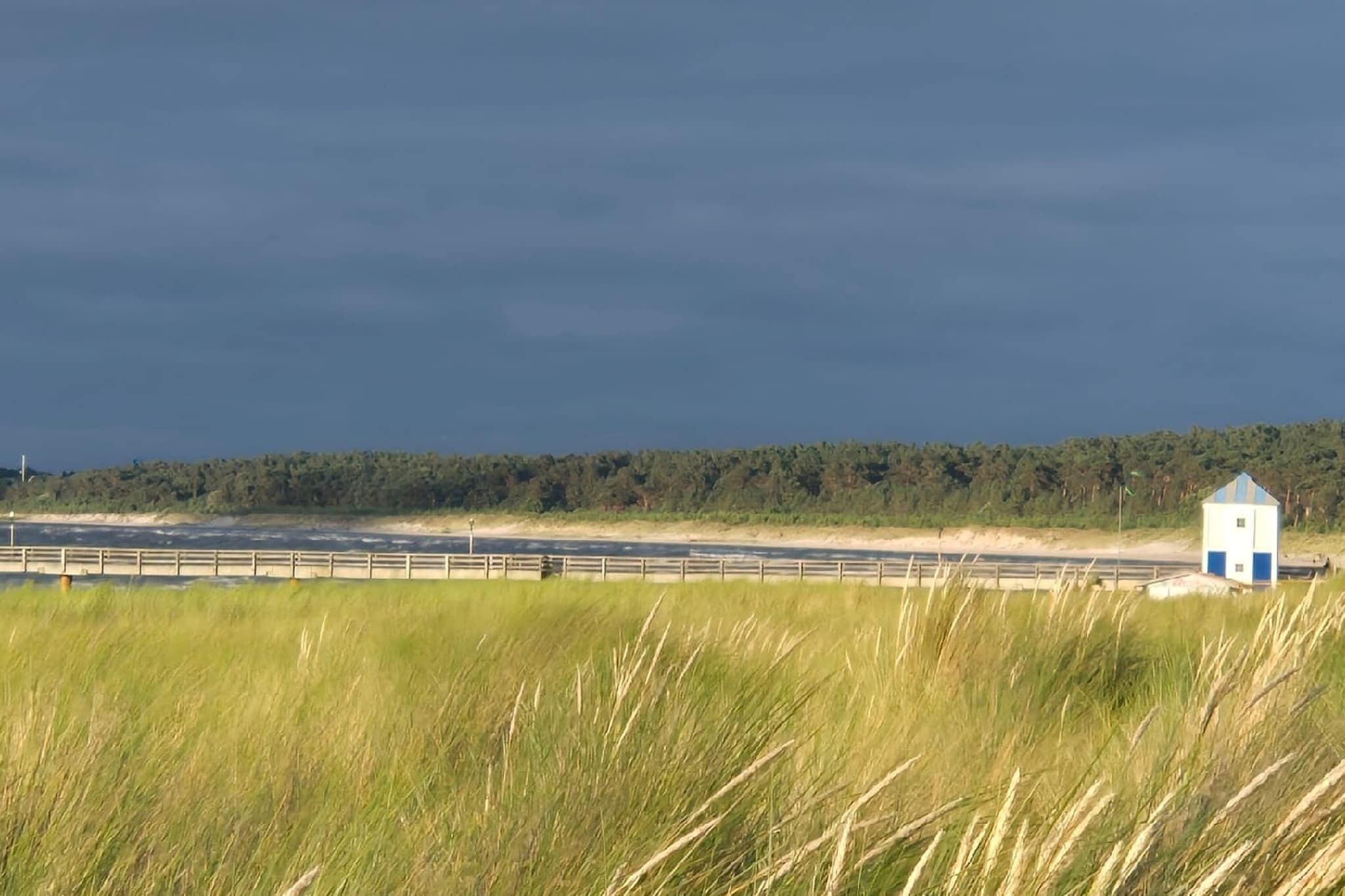 Een kustlandschap met een klein, blauw-wit gebouw, mogelijk een vuurtoren of een weerstation, nabij een zandstrand en grasduinen.