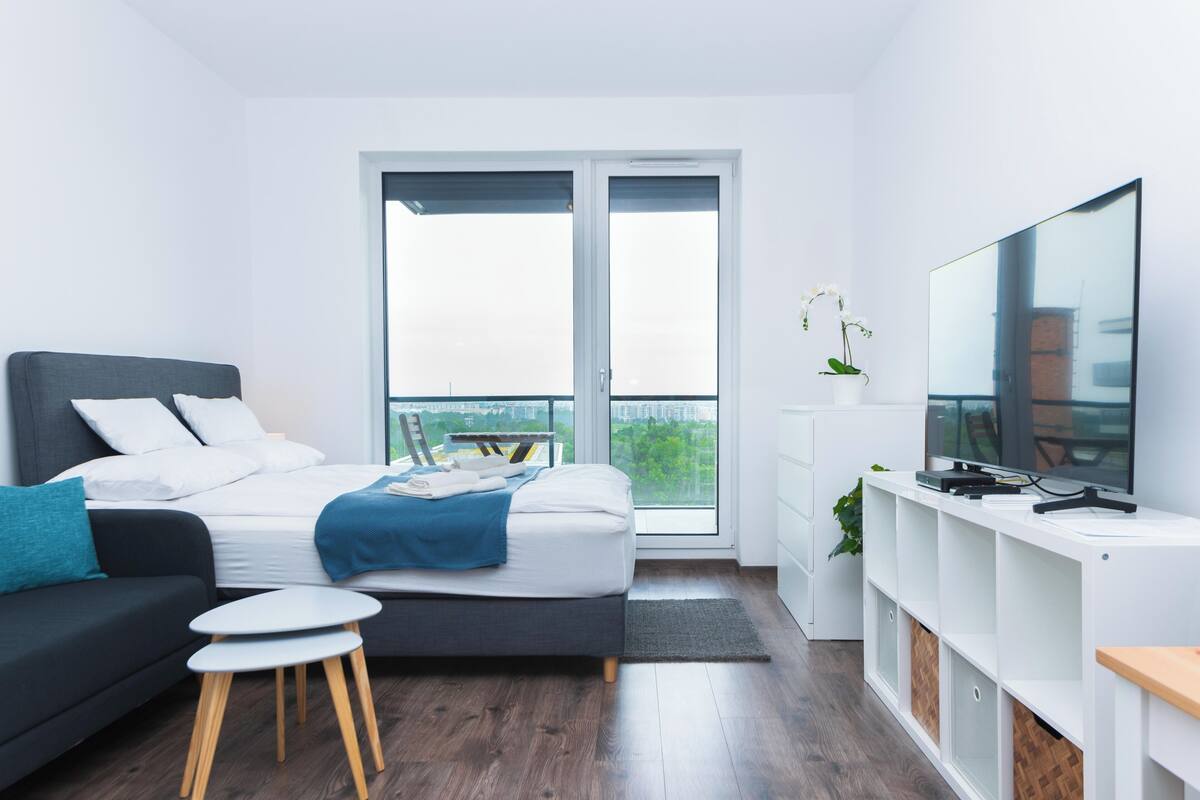 A modern bedroom with a bed, a sofa, a TV, and a balcony with a city view.
