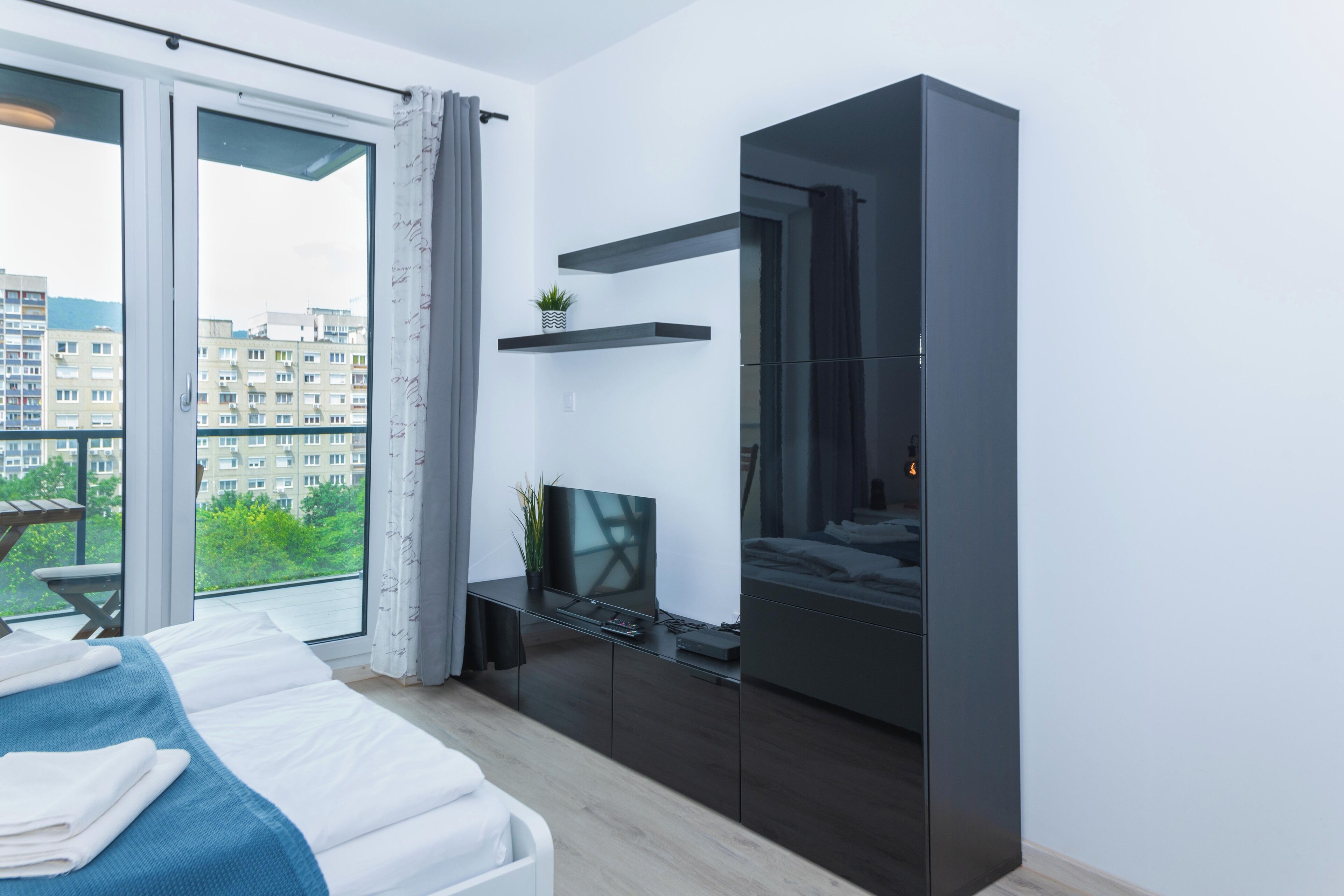 Une chambre moderne avec un lit, une télévision, une armoire sombre et un balcon donnant sur des immeubles.