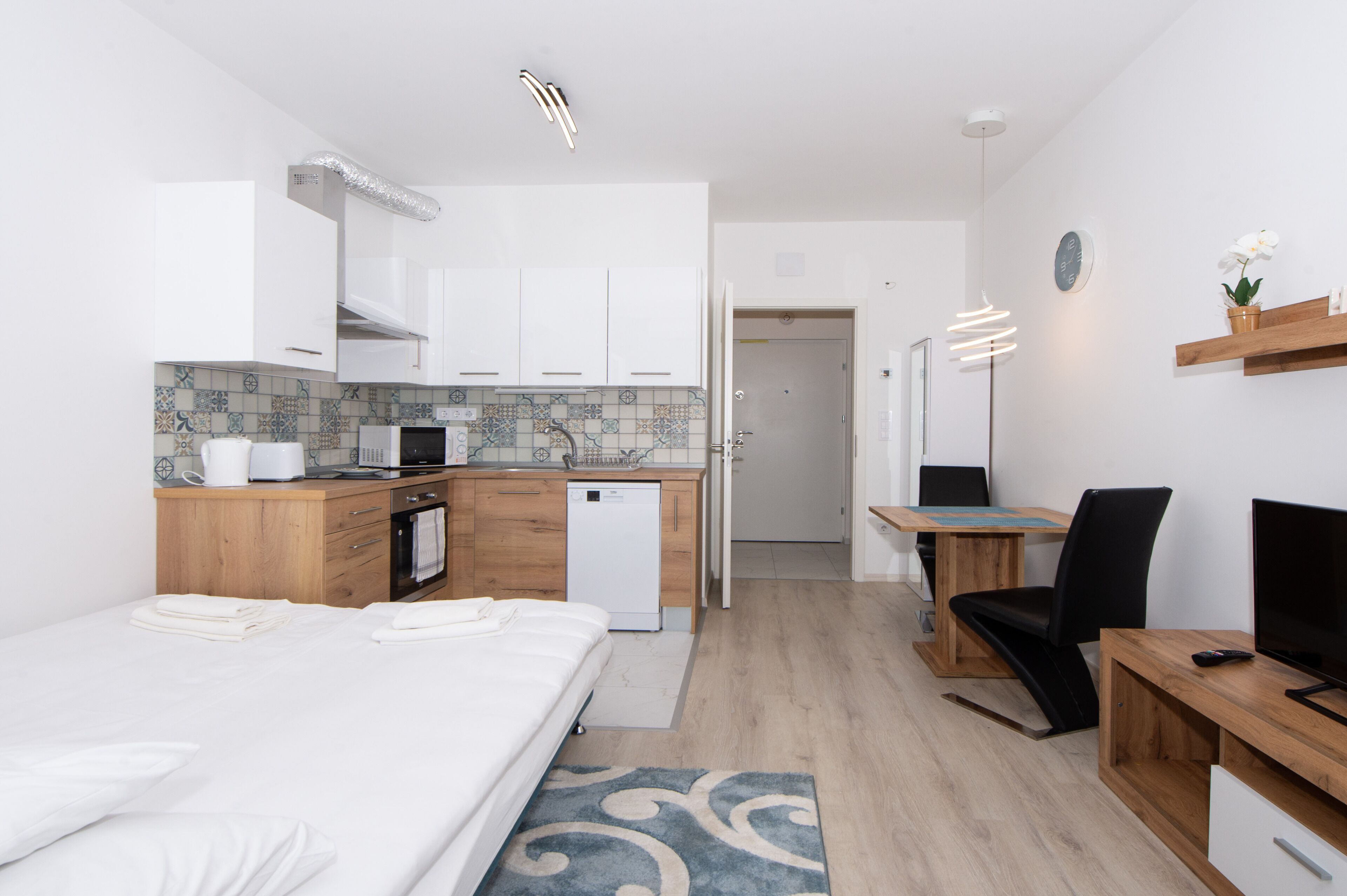 A modern hotel room with a bed, a desk, a chair, a small kitchenette, and a bathroom.