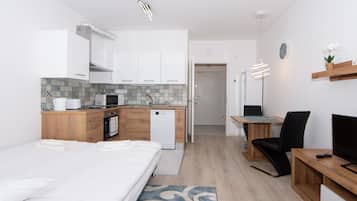 A modern hotel room with a bed, a desk, a chair, a small kitchenette, and a bathroom.