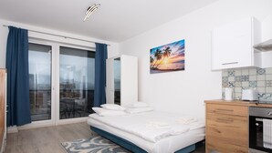A modern bedroom with a bed, a sliding glass door to a balcony, a kitchen area, and a painting of a sunset. -  WaterFront City Apartments by FirstHost (Budapest)