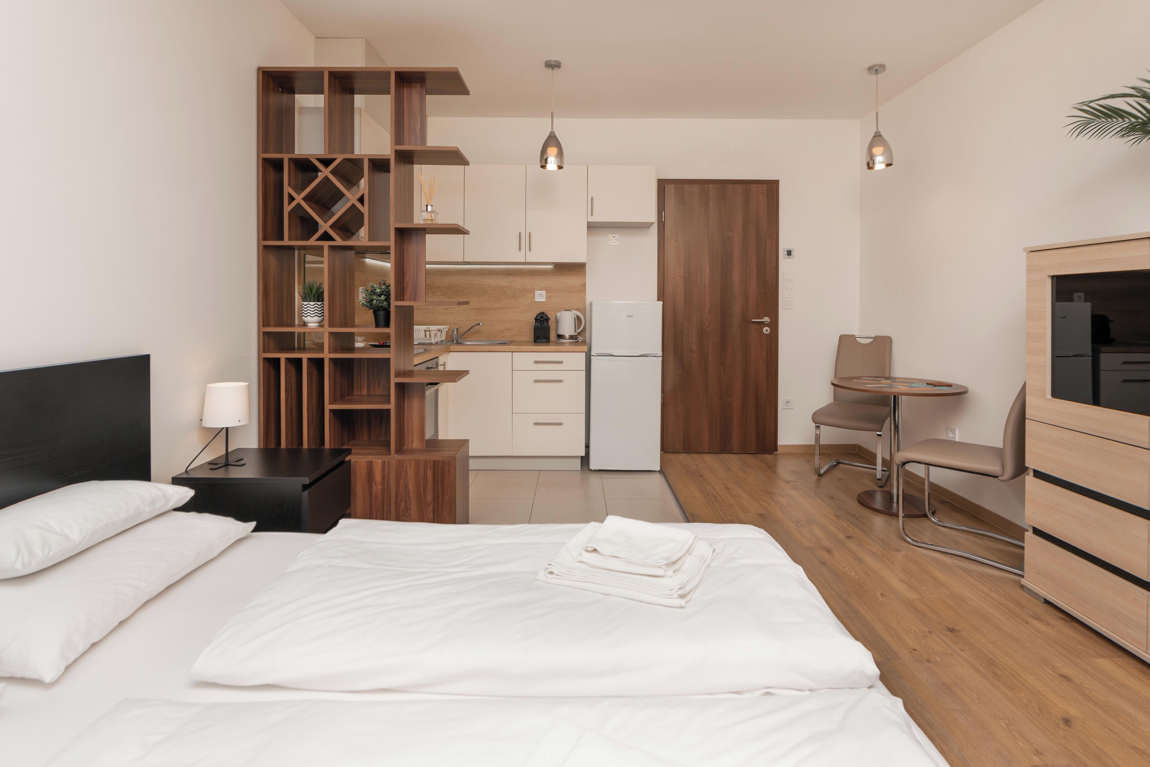 A modern hotel room with a bed, a wooden bookshelf, a small kitchen area, and a seating area with a chair and table.