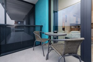 A balcony with a mesh screen, two wicker chairs, a small round table, and a view of a room with a bed and a window. -  WaterFront City Apartments by FirstHost (Budapest)