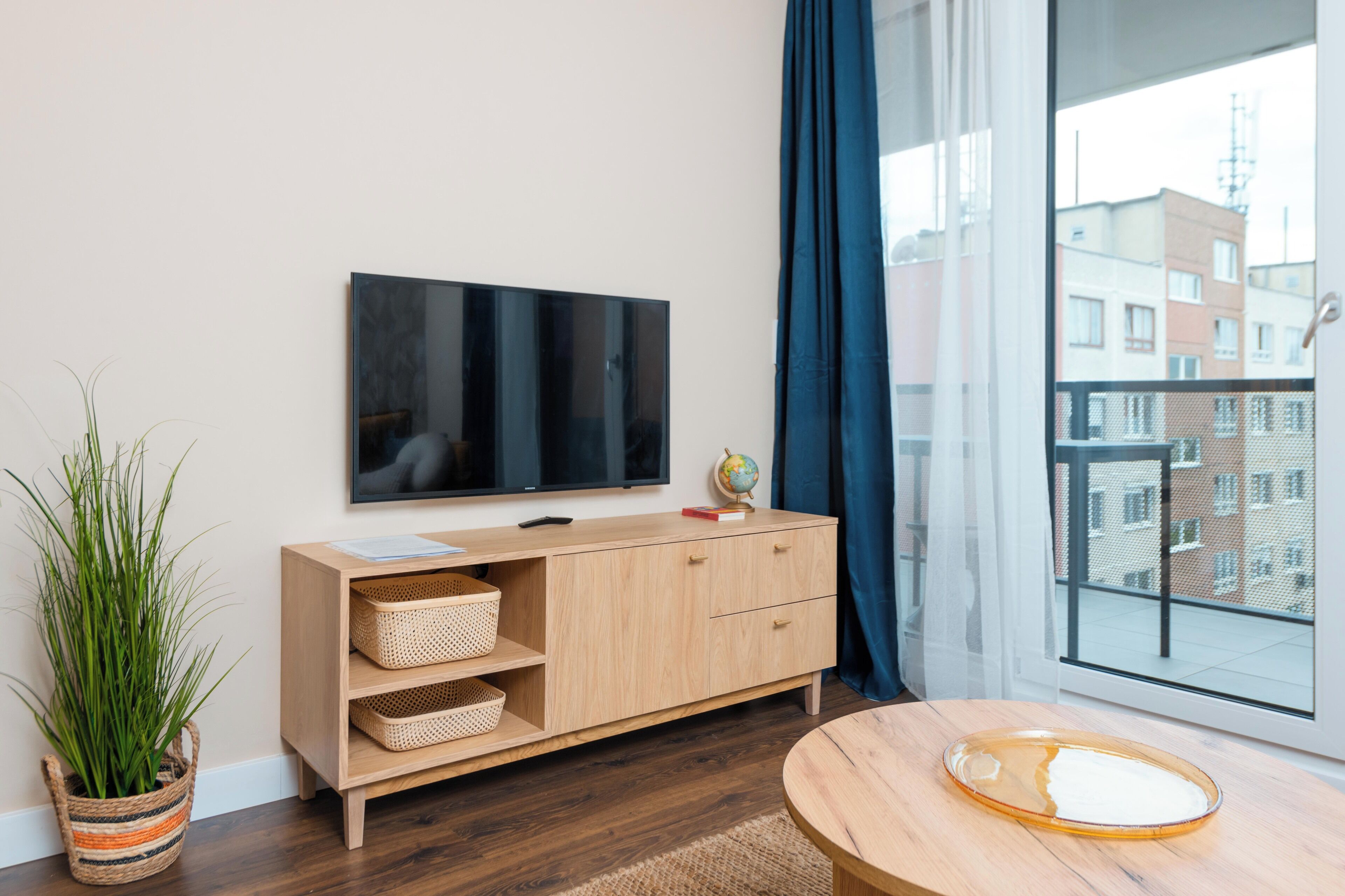 Un salon moderne avec un téléviseur à écran plat fixé au mur, un meuble en bois avec des paniers, une table ronde en bois et une grande fenêtre donnant sur des immeubles.