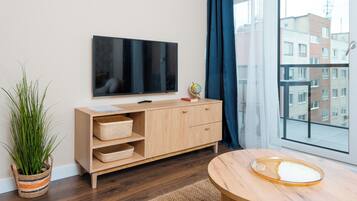 Un salon moderne avec un téléviseur à écran plat fixé au mur, un meuble en bois avec des paniers, une table ronde en bois et une grande fenêtre donnant sur des immeubles.