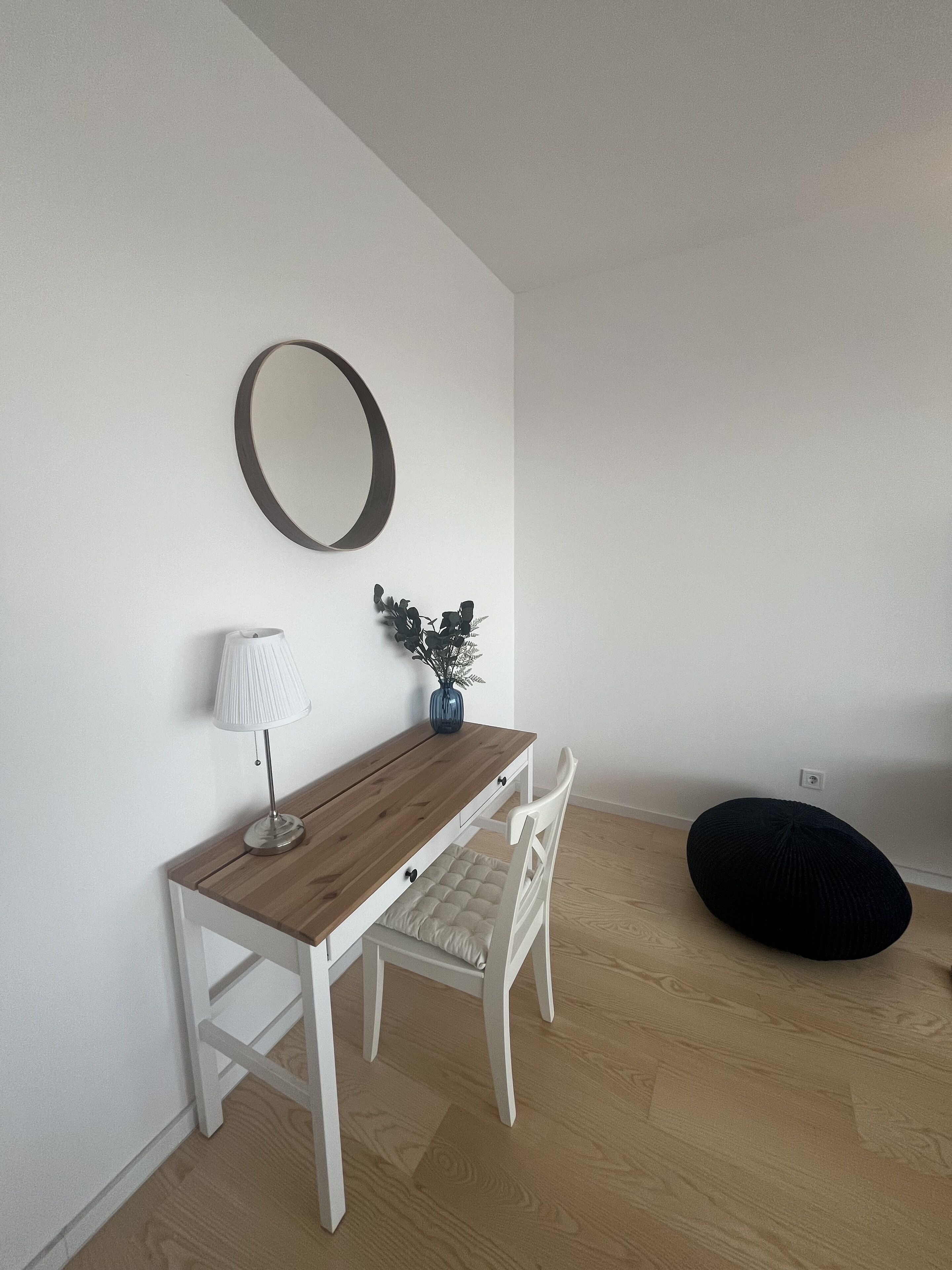 Quarto minimalista com mesa de jantar de madeira, cadeiras brancas, uma pequena planta em vaso, abajur e um puff preto.