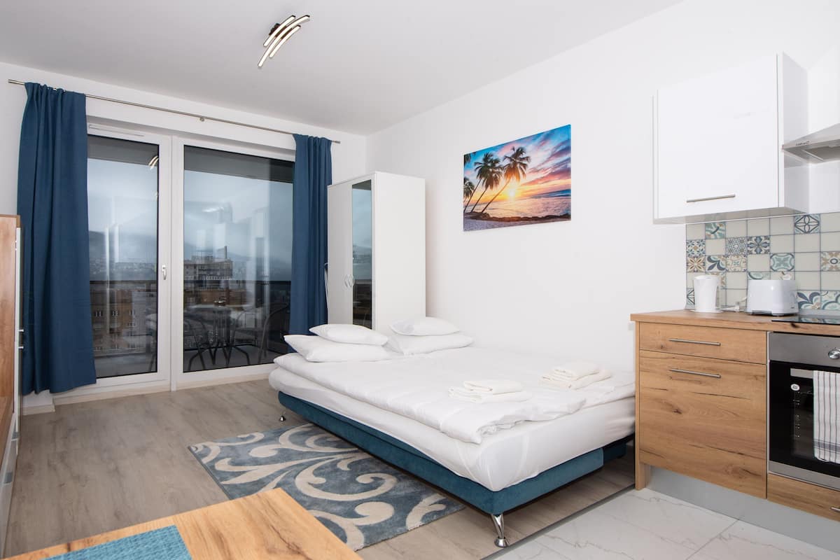 A modern bedroom with a bed, a sliding glass door to a balcony, a kitchen area, and a cityscape view.