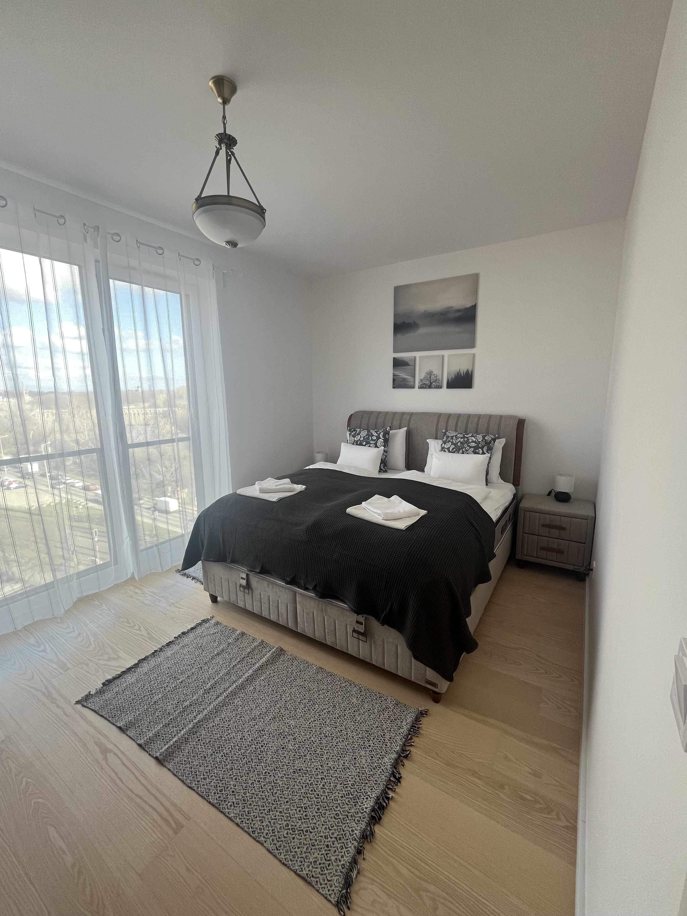 Un dormitorio con cama, mesita de noche, lámpara, ventana con cortinas y vistas al exterior.