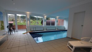 Piscina com vista para um muro de tijolos e vegetação. Há cadeiras brancas e um banco perto da piscina.